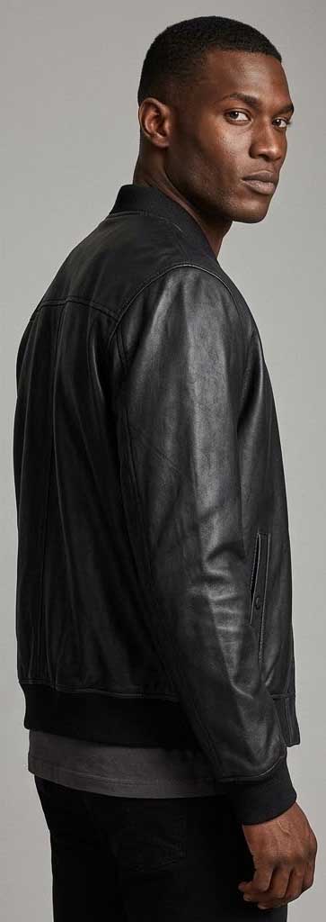 A man wearing a black leather bomber jacket, shown from a side-back angle against a neutral gray studio background.
