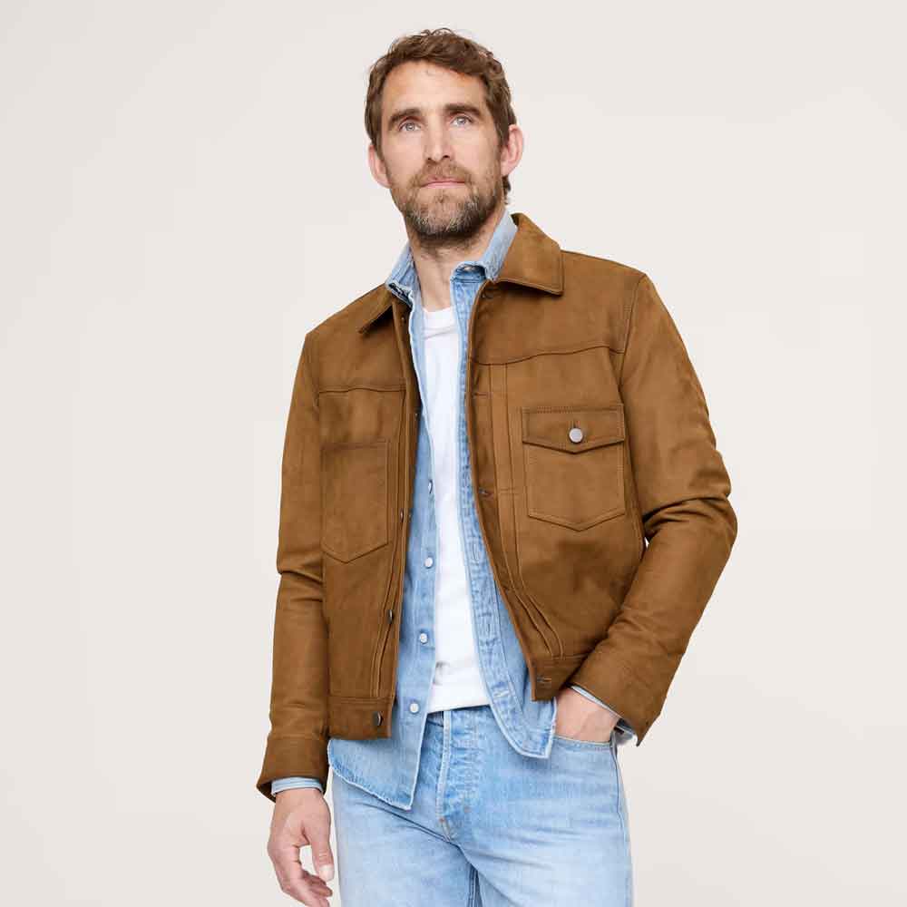 A man wearing a brown suede jacket over a light blue denim shirt and white t-shirt, paired with light blue jeans, photographed against a solid off-white background.