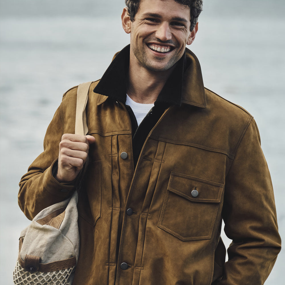 A smiling man wearing a brown suede-like jacket over a dark shirt and white undershirt, carrying a beige shoulder bag against a soft, blurred outdoor background.