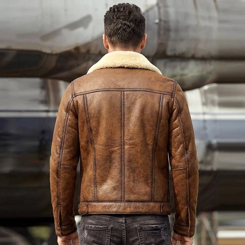 A man viewed from behind wearing a brown leather aviator-style jacket with a cream shearling collar, standing in front of a blurred aircraft.