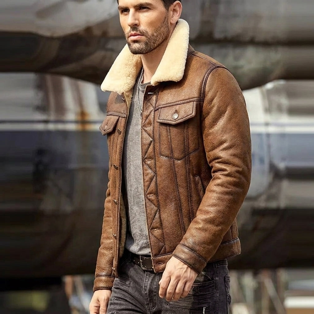 A man wearing a brown leather aviator-style jacket with a cream shearling collar, layered over a gray quilted shirt and dark jeans, standing in front of a blurred aircraft.