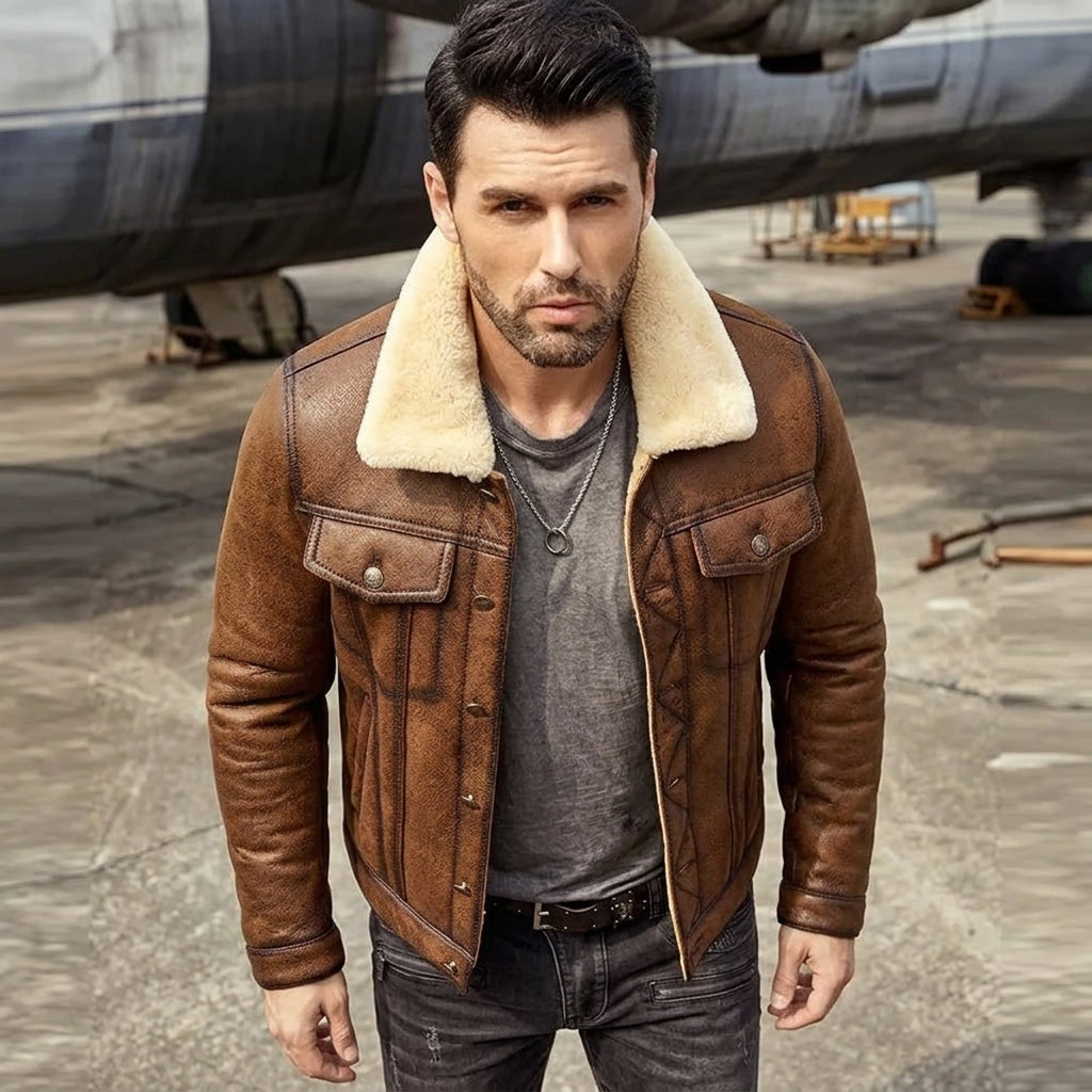 A man wearing a brown leather jacket with a cream shearling collar, layered over a gray t-shirt and dark jeans, standing in front of an industrial aircraft on a concrete tarmac.