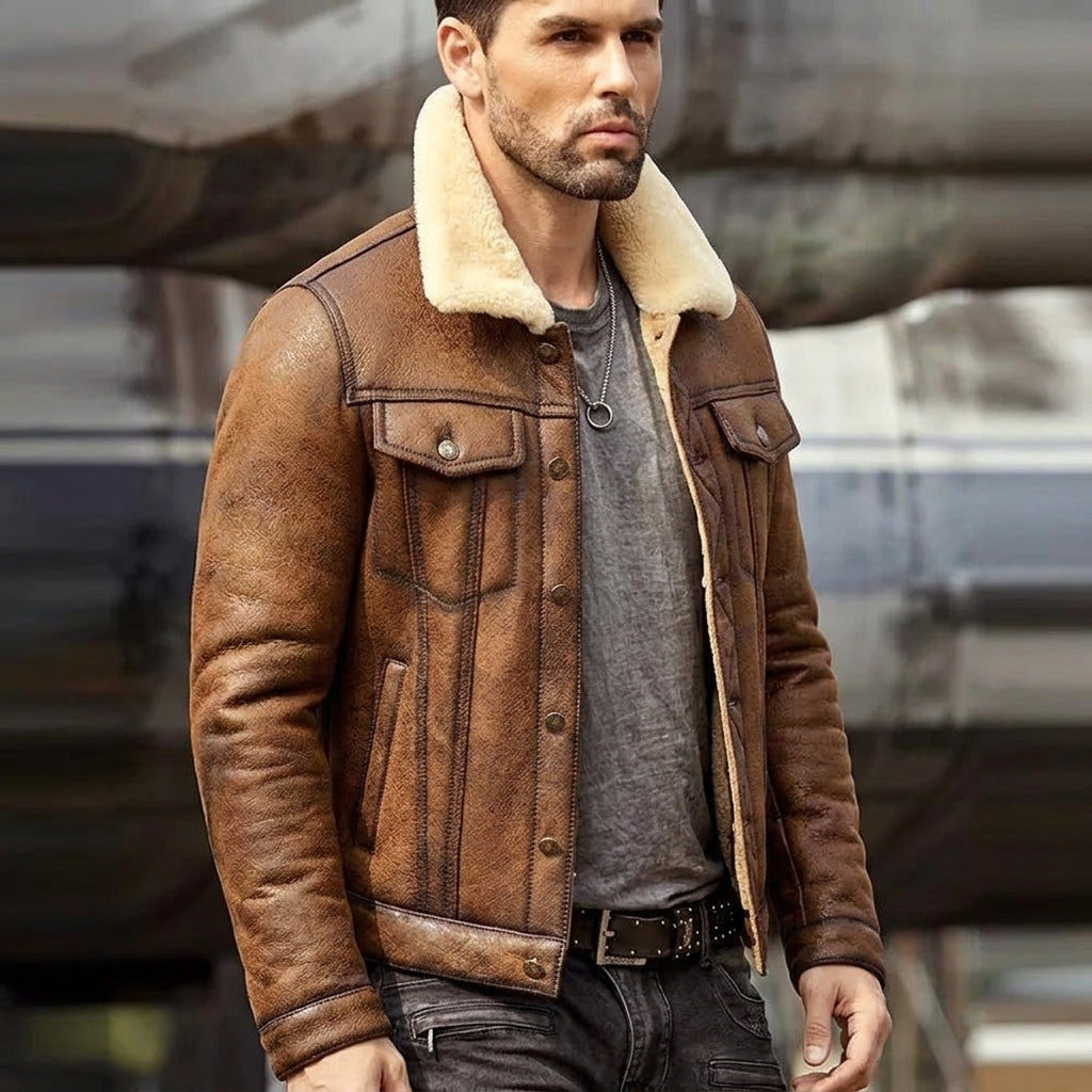 A man wearing a brown leather aviator-style jacket with a cream shearling collar, layered over a gray t-shirt and dark jeans, posed in front of a blurred industrial background.