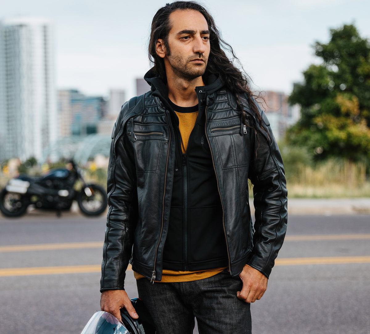 Man wearing a black leather jacket standing on a street with a motorcycle in the background.