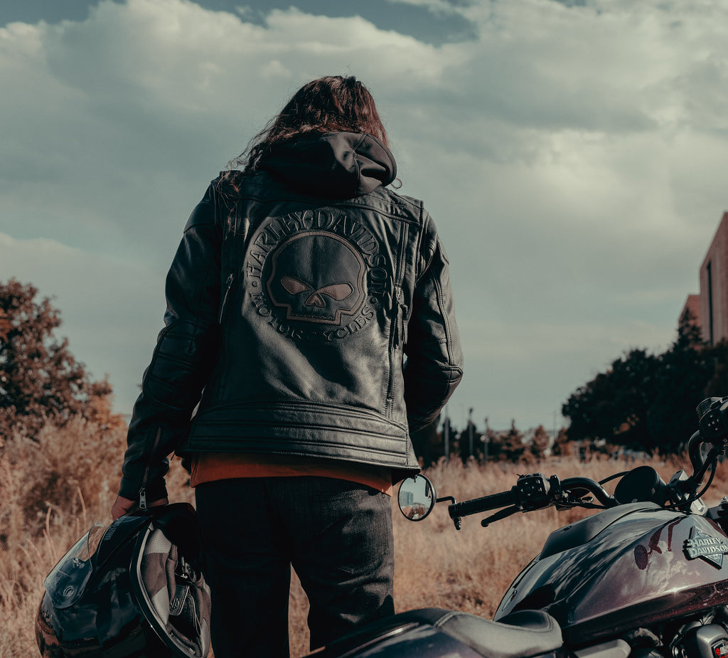 Person wearing a leather jacket with a skull emblem standing next to a motorcycle in an open field.