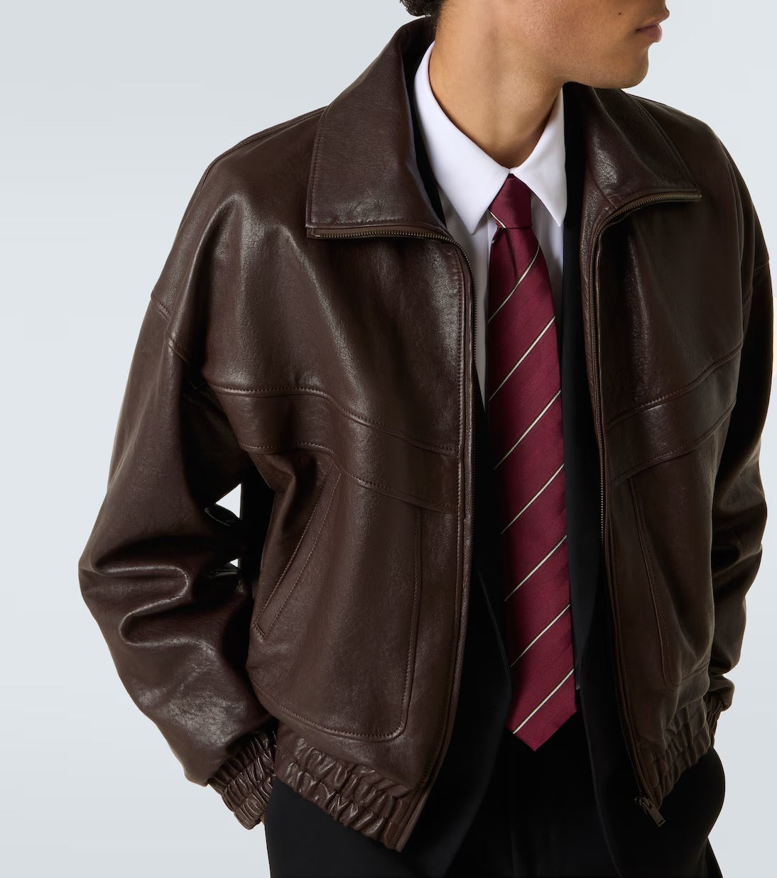 A person wearing a dark brown leather jacket over a white collared shirt and a burgundy striped tie, shown from the neck down against a light gray studio background.