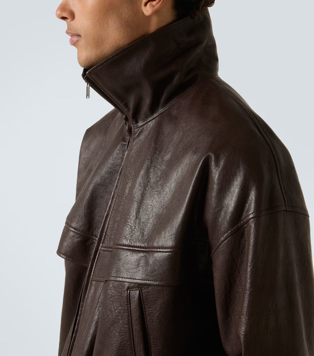 A close-up side profile of a model wearing a dark brown leather jacket with a high collar and zippered front, set against a light gray studio background.