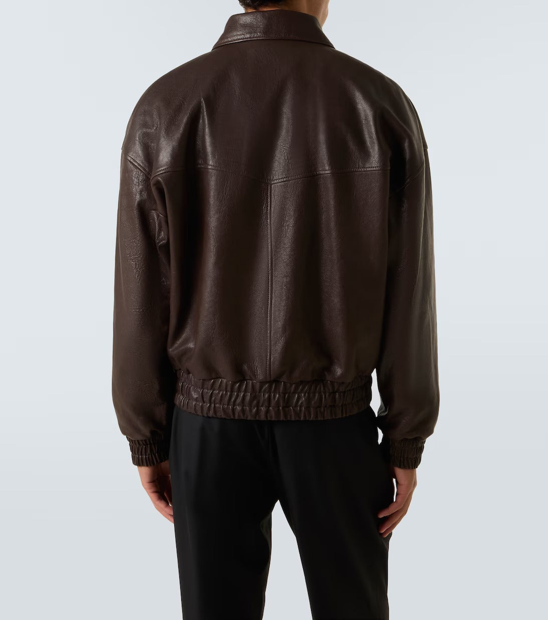 A person shown from the back wearing a dark brown leather jacket with a gathered elastic hem and cuffs, paired with black pants, against a light gray studio background.