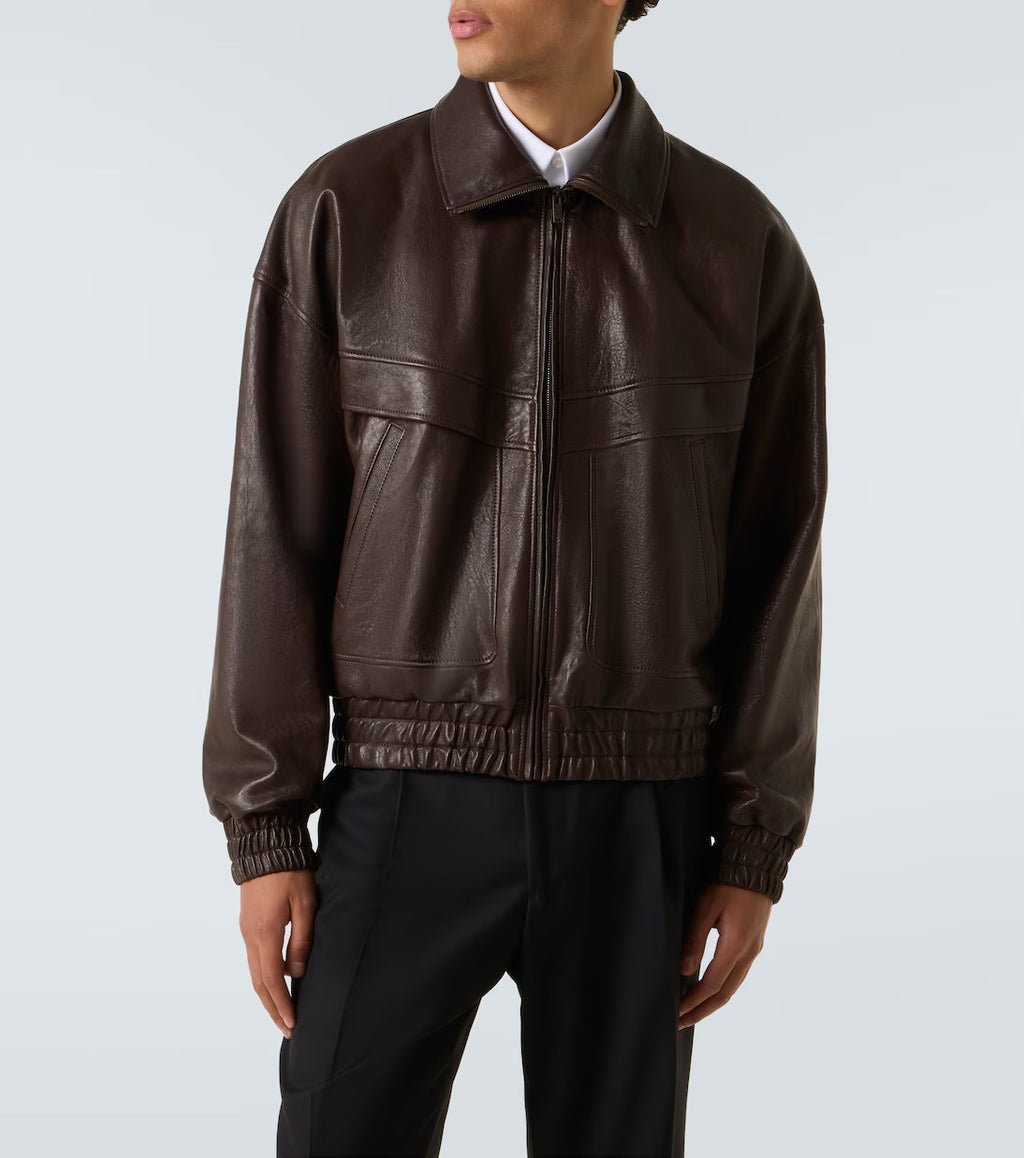 A model wearing a dark brown leather jacket with a front zipper, collar, and elasticized hem, paired with black trousers against a light gray studio background.