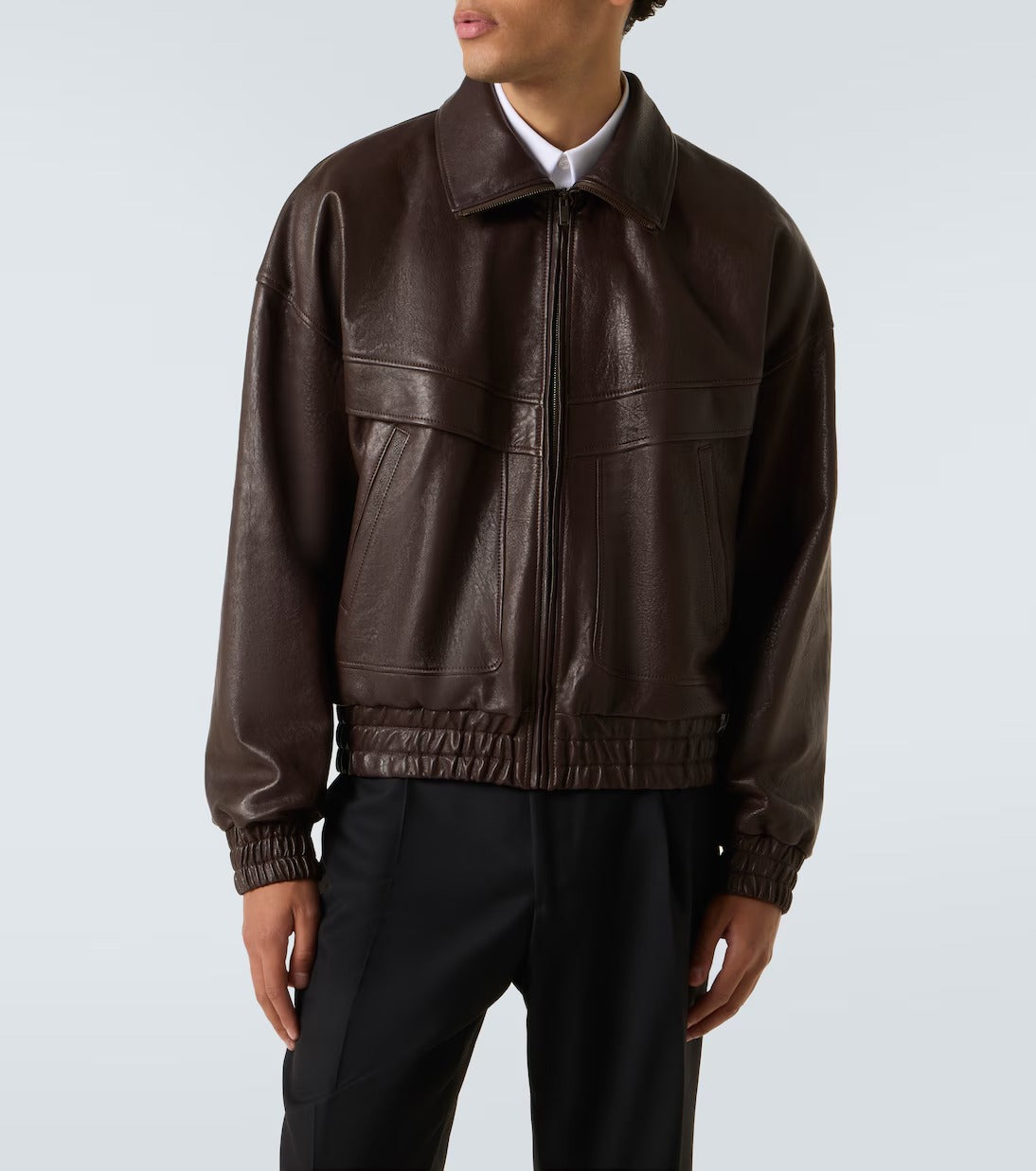 A model wearing a dark brown leather jacket with a front zipper, collar, and elasticized hem, paired with black trousers against a light gray studio background.