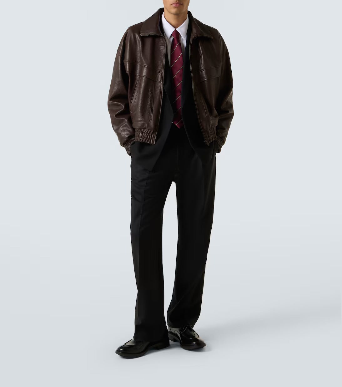 A man wearing a dark brown leather jacket over a black suit, white dress shirt, and a red striped tie, standing against a light gray studio background.