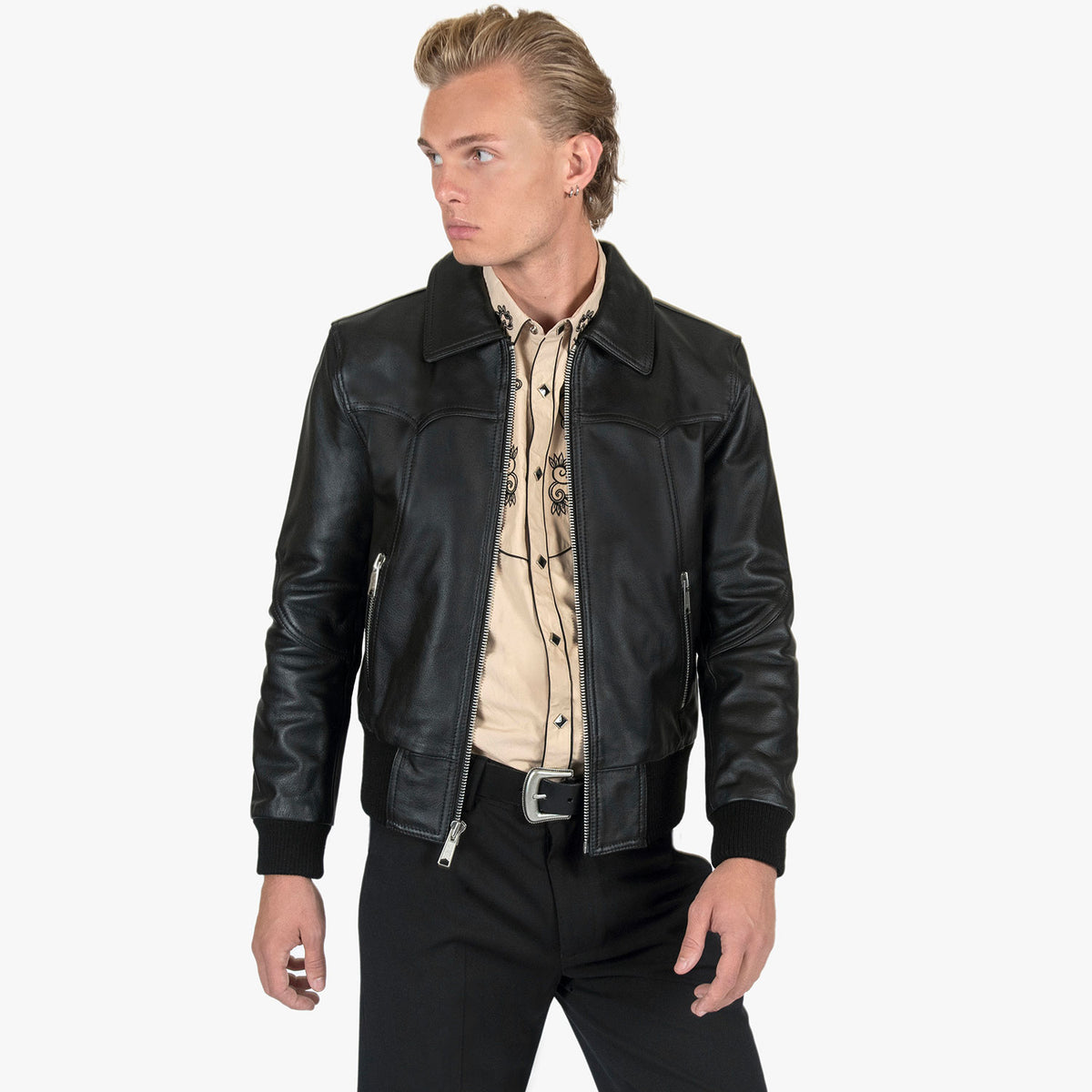 A man wearing a black leather motorcycle jacket over a beige western-style shirt and black pants, photographed against a plain light gray studio background.