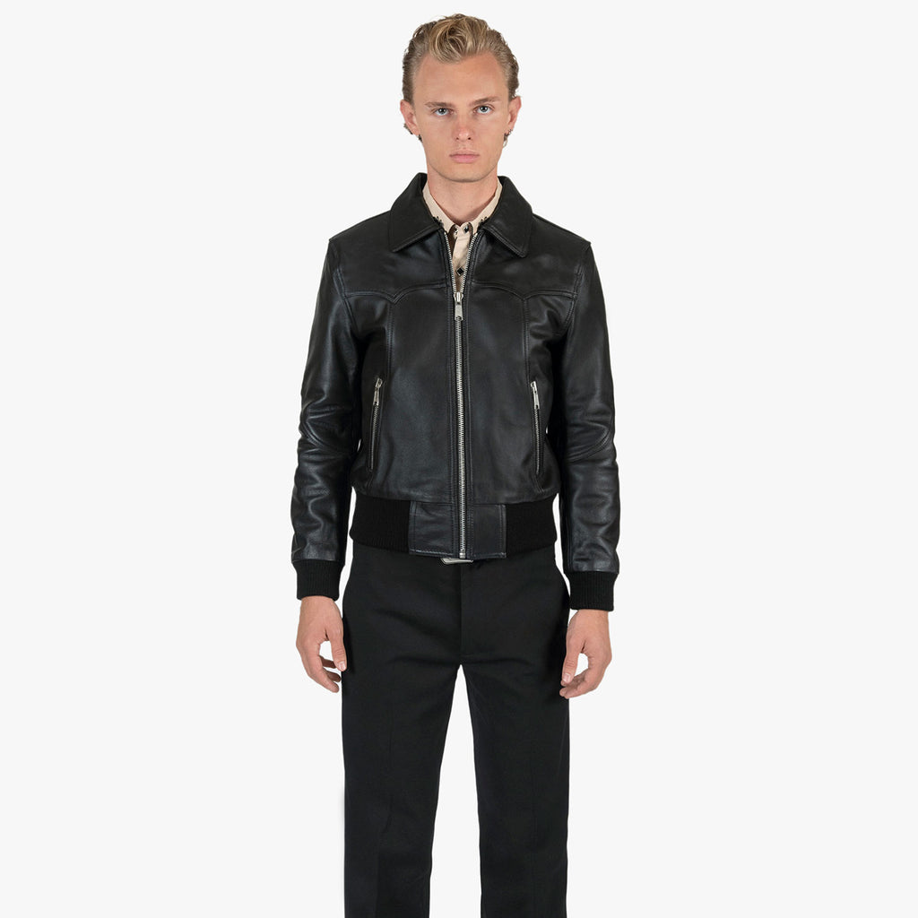 A male model wearing a black leather biker jacket with silver zippers, paired with black trousers and a light-colored shirt, standing against a plain white background.