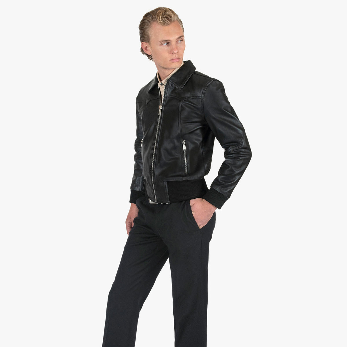 A man wearing a black leather jacket with silver zippers and black trousers, standing against a light gray studio background.