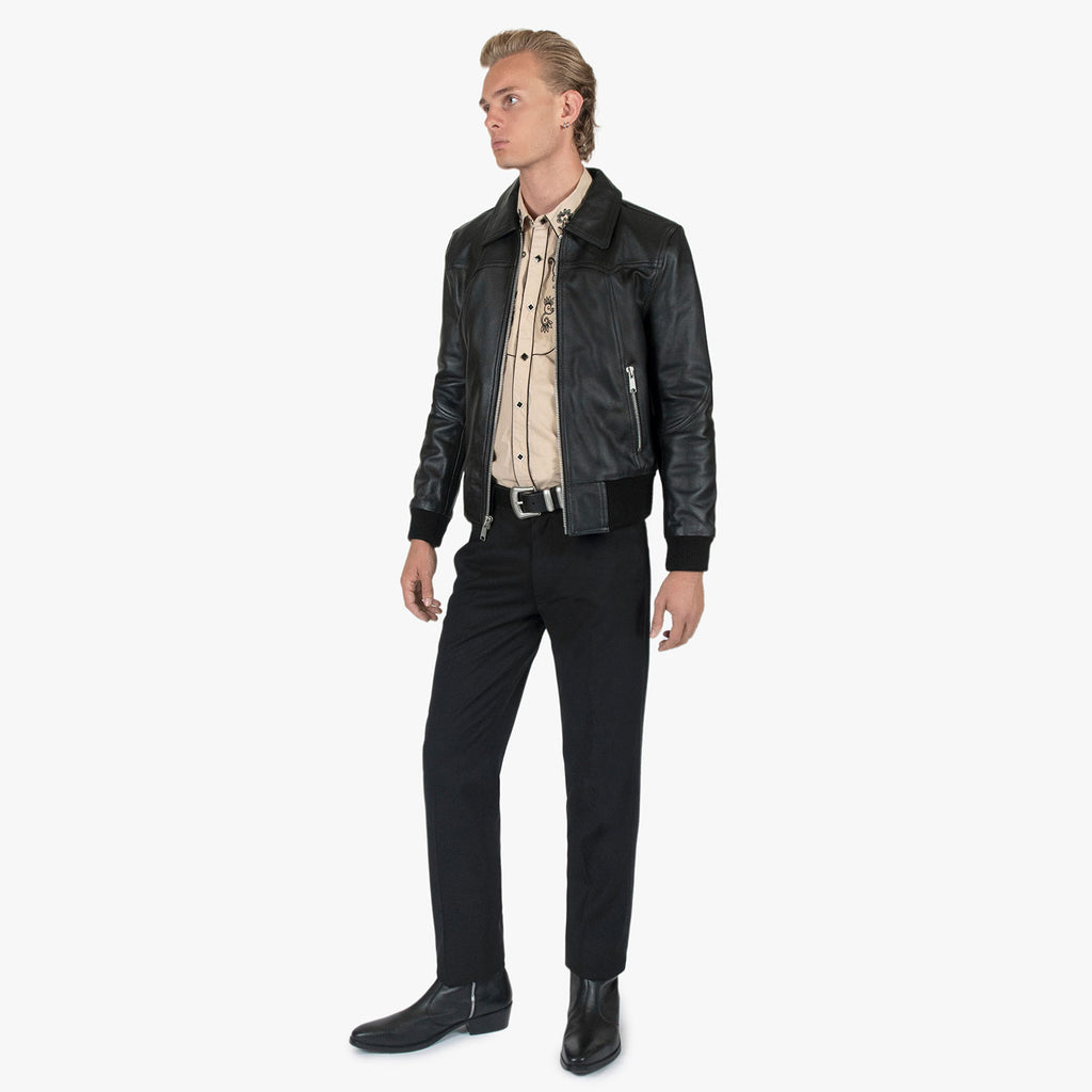 A man wearing a black leather motorcycle jacket over a beige patterned button-down shirt, black trousers, and black boots, standing against a light gray studio background.