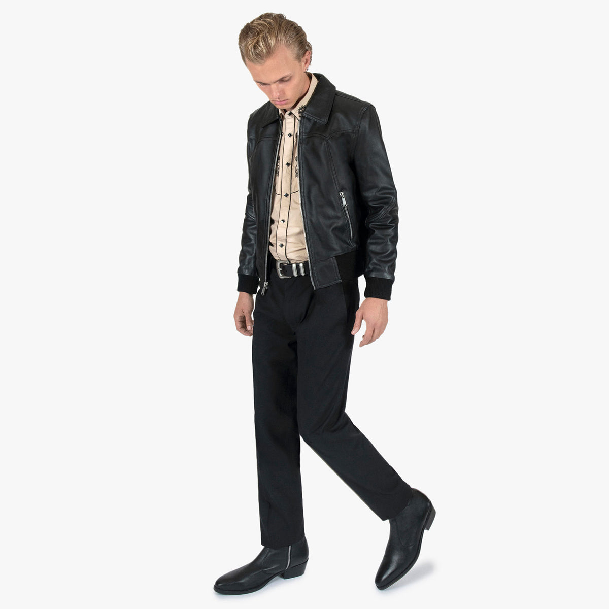 A man modeling a black leather jacket, beige patterned shirt, black trousers, and black leather boots against a light gray studio background.
