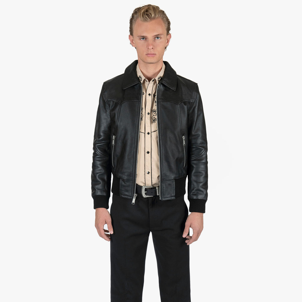 A male model wearing a black leather motorcycle jacket over a beige western-style shirt, paired with black trousers, against a plain light gray background.