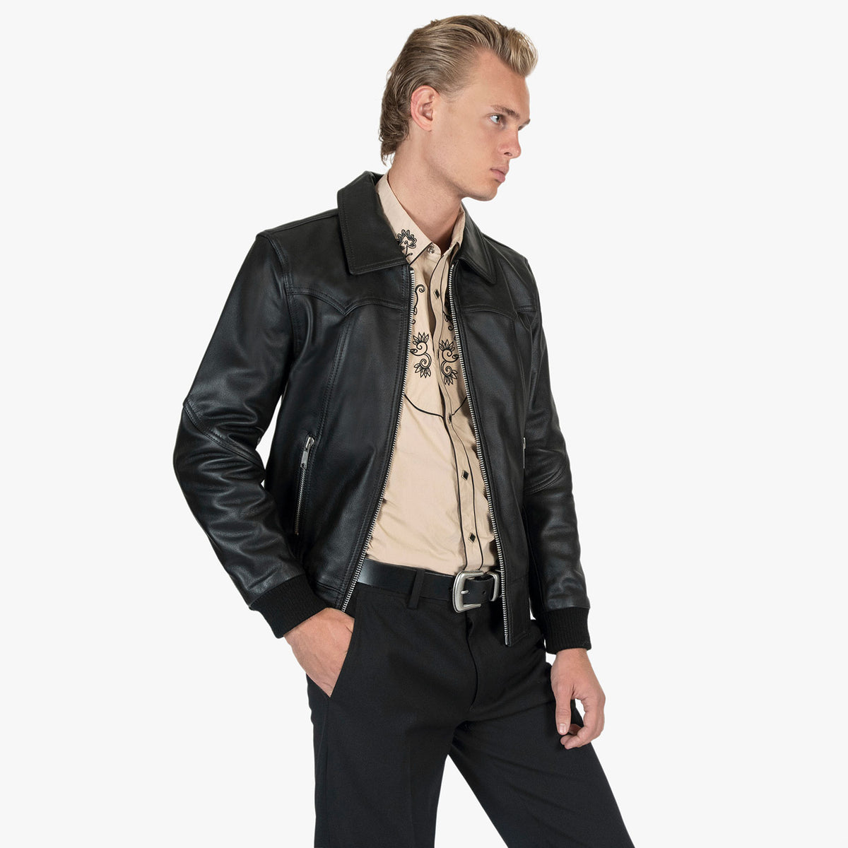 A man wearing a black leather jacket over a beige embroidered shirt and black trousers, photographed against a plain light gray studio background.