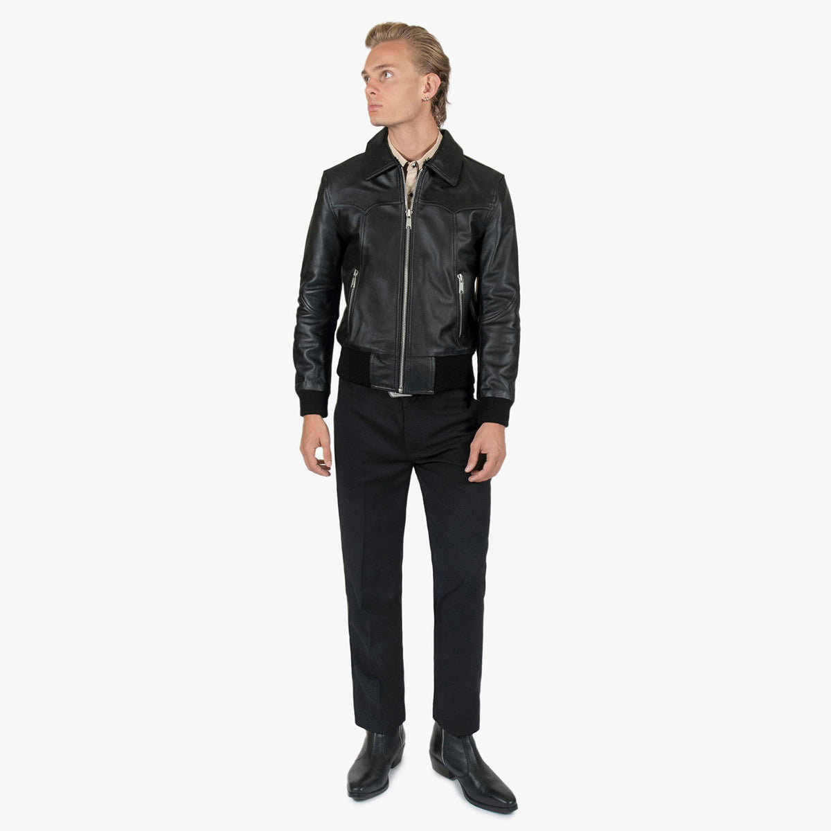 A man wearing a black leather biker jacket with a silver zipper, black trousers, and black leather boots, standing against a plain white studio background.