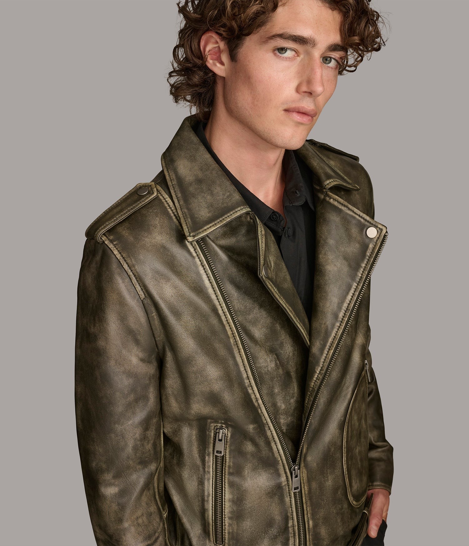 A man wearing a distressed olive-brown leather motorcycle jacket over a black shirt, photographed against a solid gray studio background.