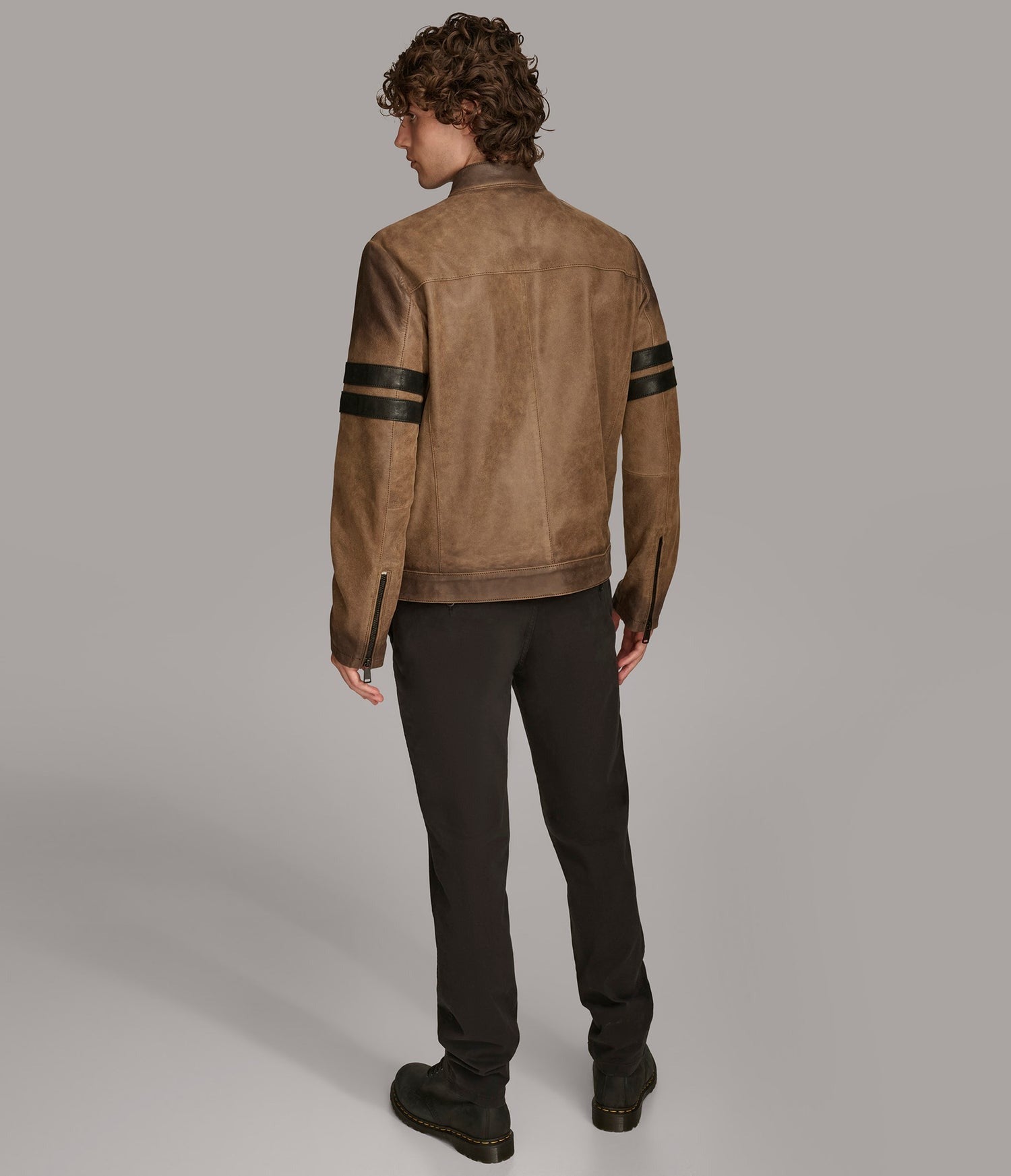 A man shown from the back wearing a brown suede jacket with black stripe details on the sleeves, paired with black pants and boots, against a solid gray studio background.