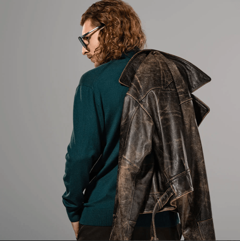 A model with curly brown hair and glasses, wearing a dark teal knit sweater and a distressed dark brown leather jacket, photographed from the back against a solid gray studio background.