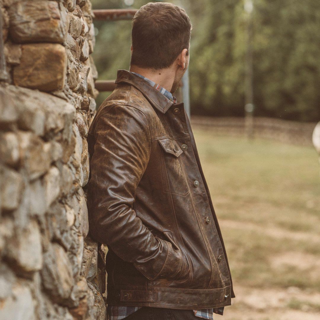 Man wearing a brown leather jacket leaning against a stone wall outdoors.