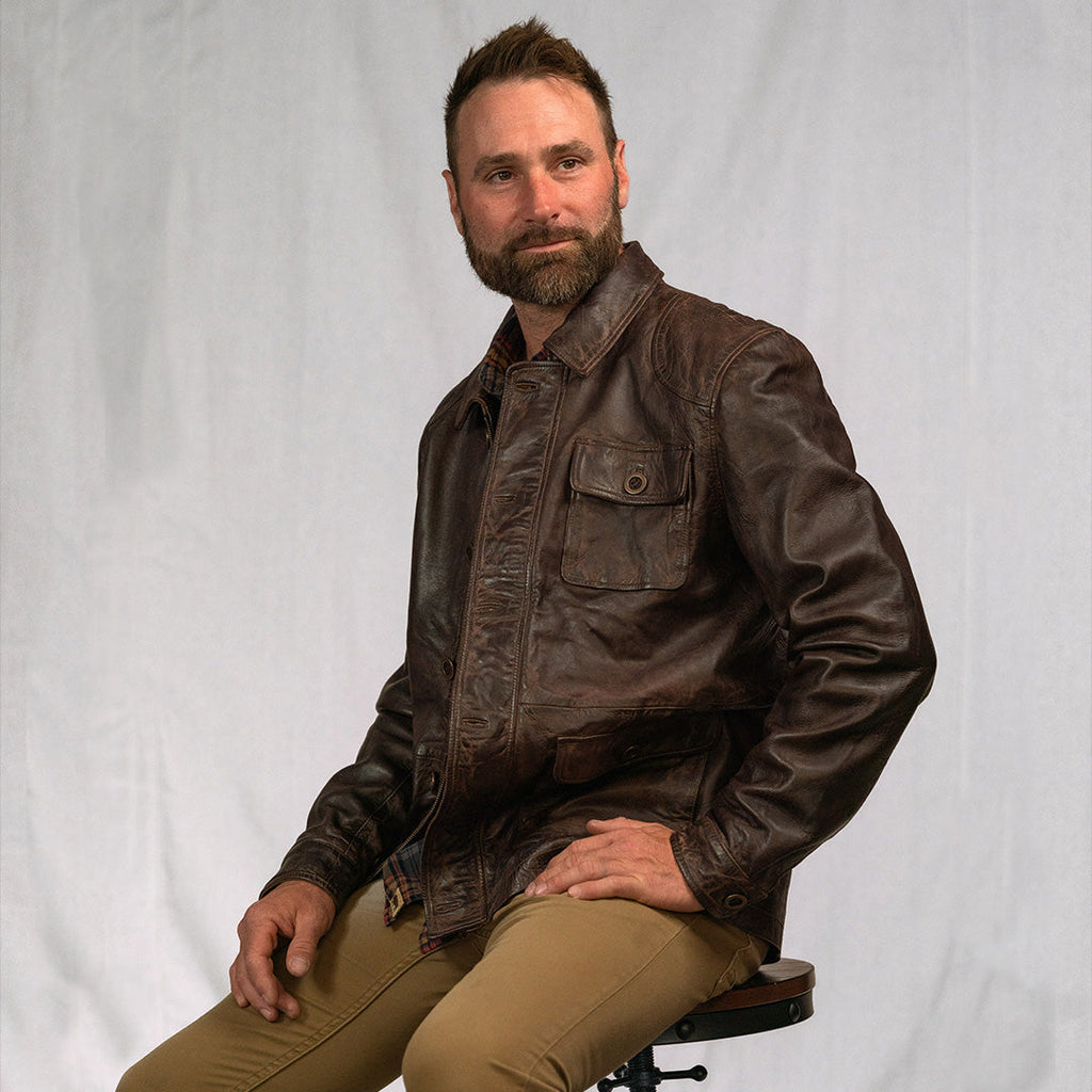 Man wearing a brown leather jacket sitting on a chair against a white background