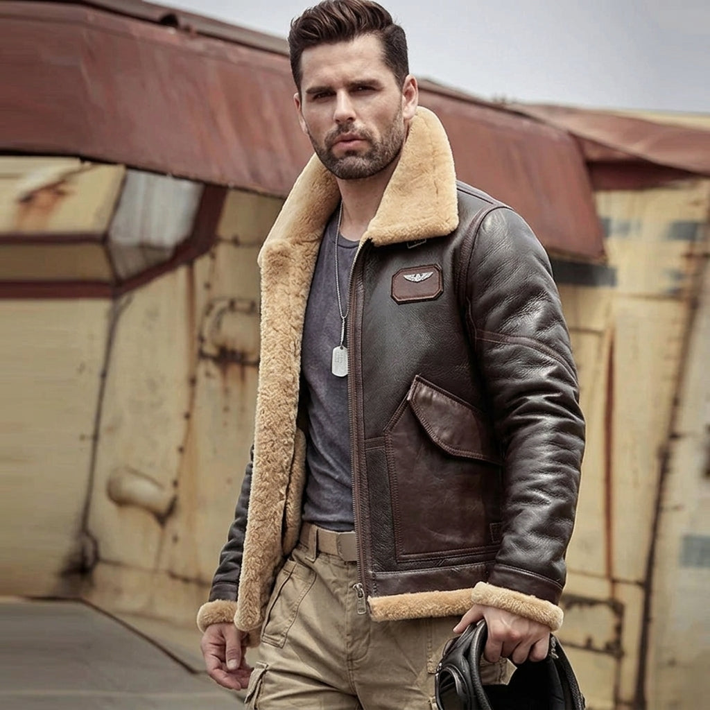 A man wearing a brown leather aviator-style jacket with a thick beige shearling collar, standing outdoors in front of a weathered industrial building.