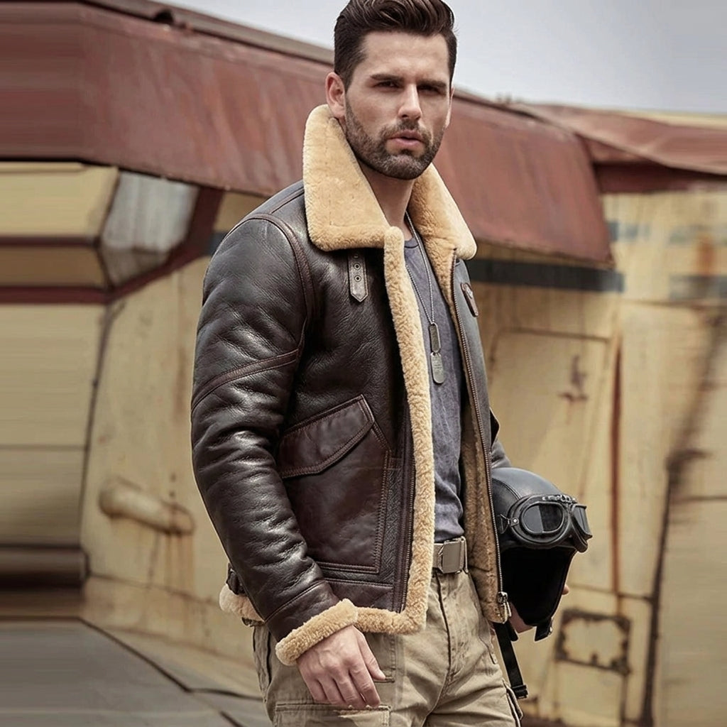 A man wearing a brown leather aviator jacket with a beige shearling lining, standing outdoors in front of a rustic building while holding a black helmet with goggles.