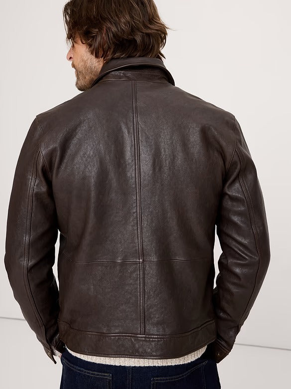 A man viewed from behind wearing a dark brown leather jacket, paired with a cream knit sweater and dark blue jeans, against a plain off-white background.