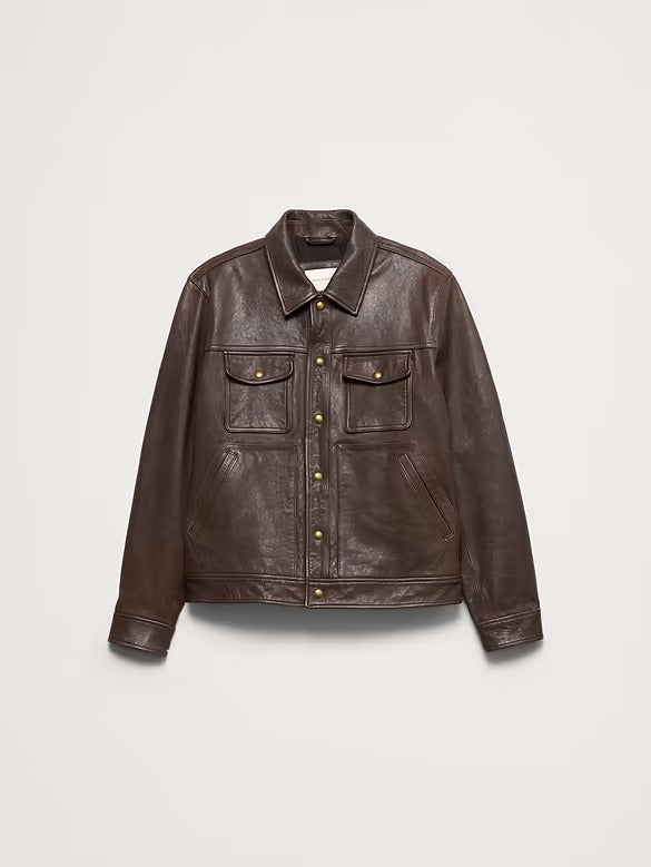 A dark brown leather jacket with a button-up front, two chest flap pockets, and gold-tone hardware, displayed flat against a light gray background.