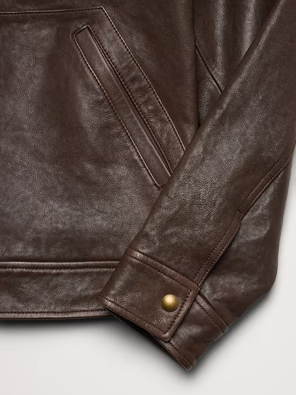 Close-up of a dark brown leather jacket showing the textured leather material, a diagonal welt pocket, and a brass snap-button cuff on a light gray background.