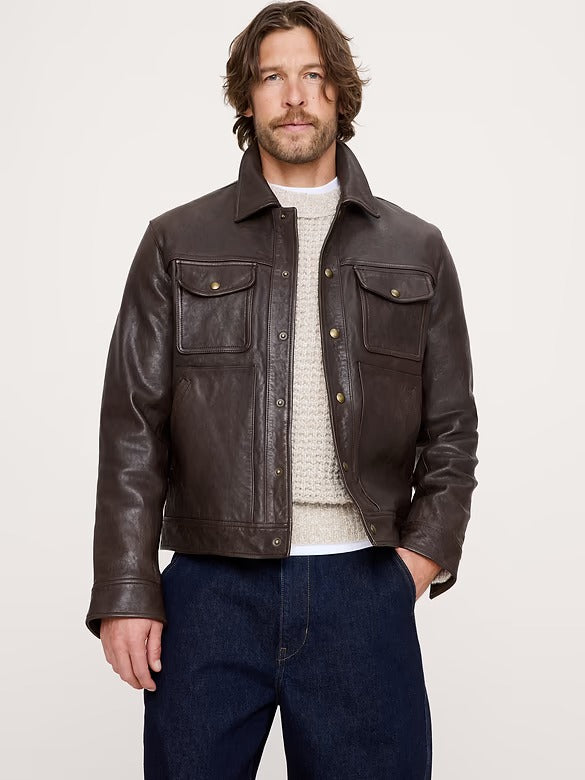 A man wearing a dark brown leather jacket over a cream knit sweater and dark blue jeans, standing against a light gray studio background.