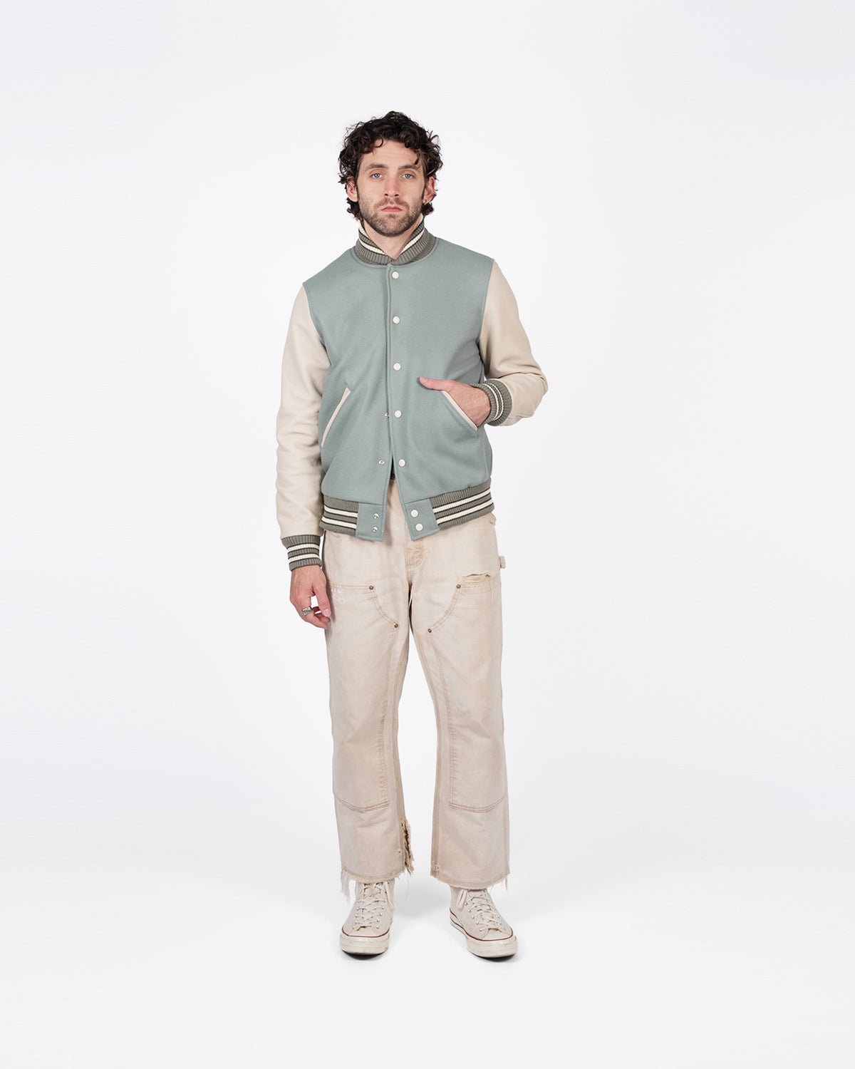Man wearing a green varsity jacket over a beige sweater with matching pants on a white background