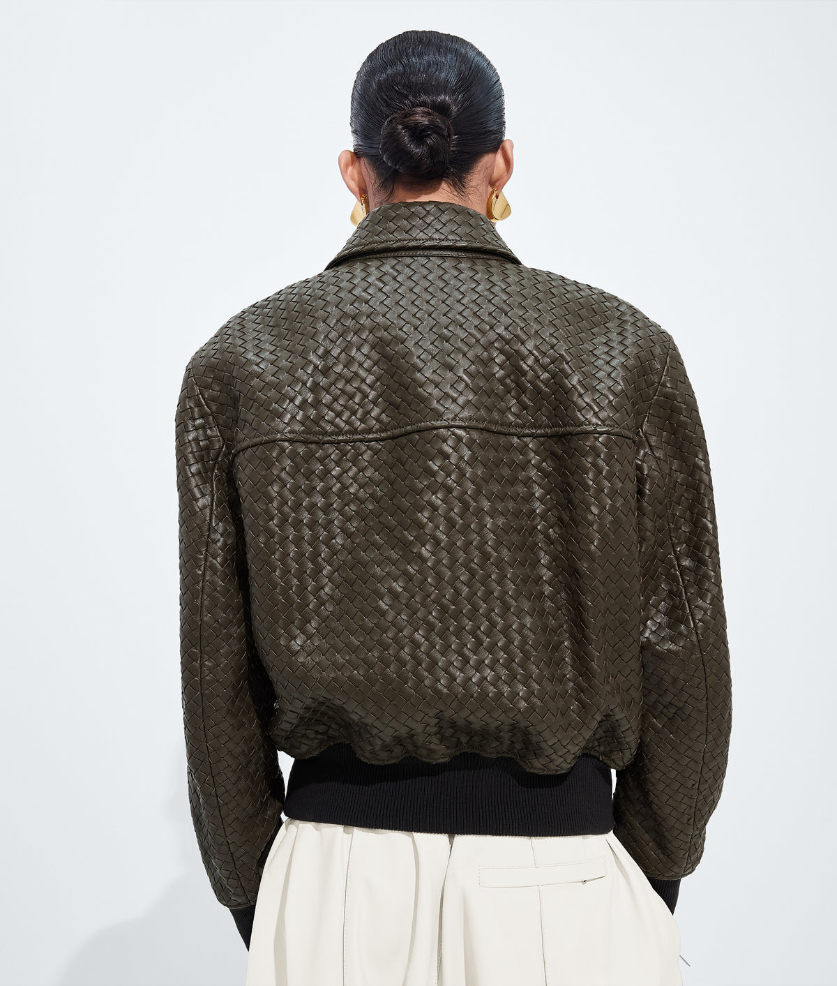 A person shown from the back wearing a dark olive-brown woven leather jacket with a textured basketweave pattern, paired with a black top and cream-colored trousers, against a plain white background.