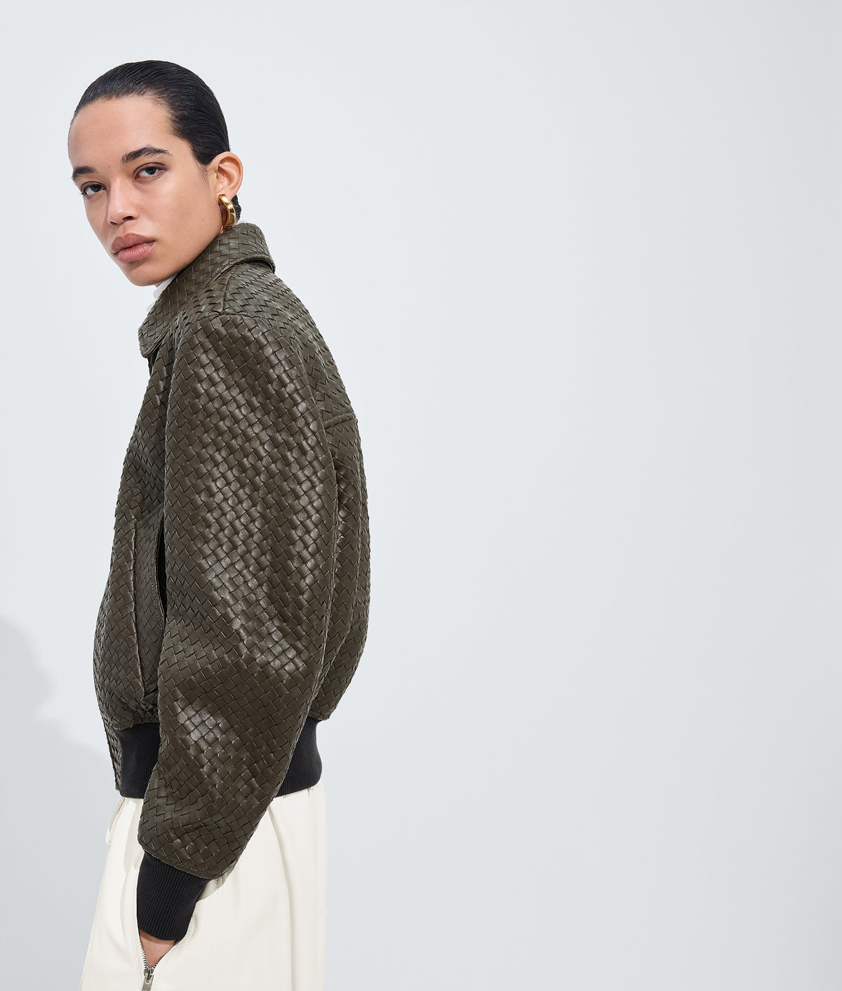 A model shown from the side profile wearing a textured olive green woven jacket with black ribbed trim, paired with off-white trousers, against a light gray studio background.
