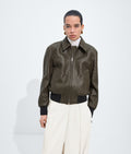 A model stands against a light gray studio background wearing a dark olive-brown textured zip-up jacket with black ribbed trim, paired with cream wide-leg trousers and a white turtleneck.