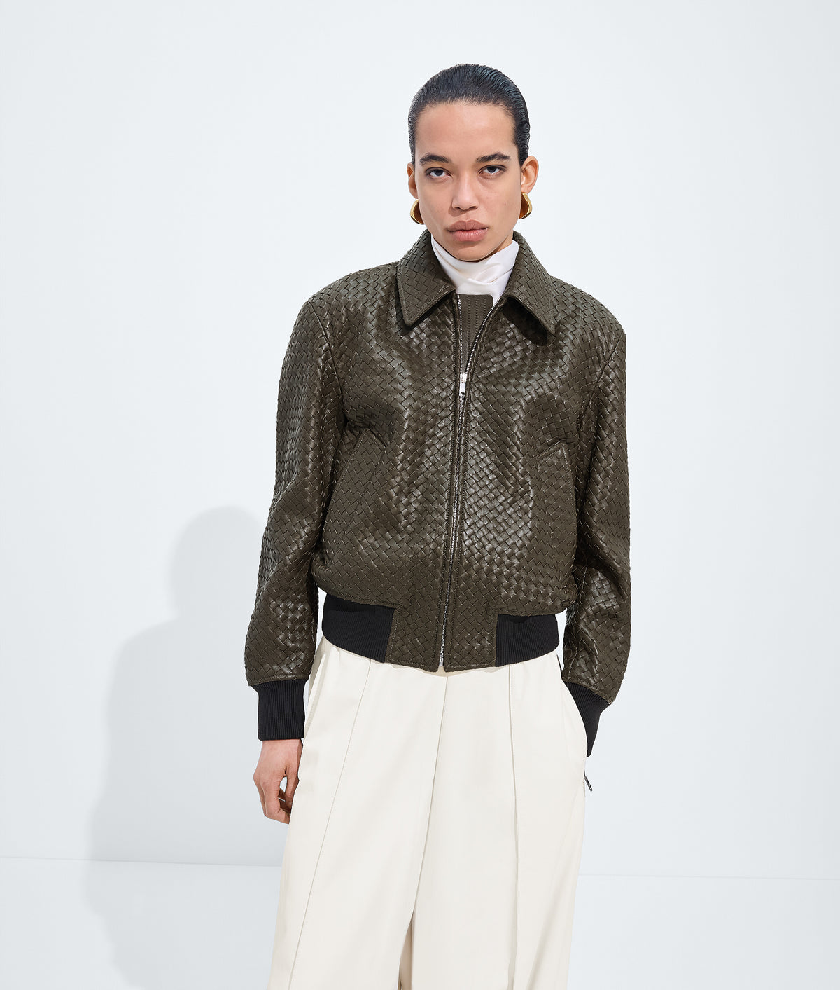 A model stands against a light gray studio background wearing a dark olive-brown textured zip-up jacket with black ribbed trim, paired with cream wide-leg trousers and a white turtleneck.