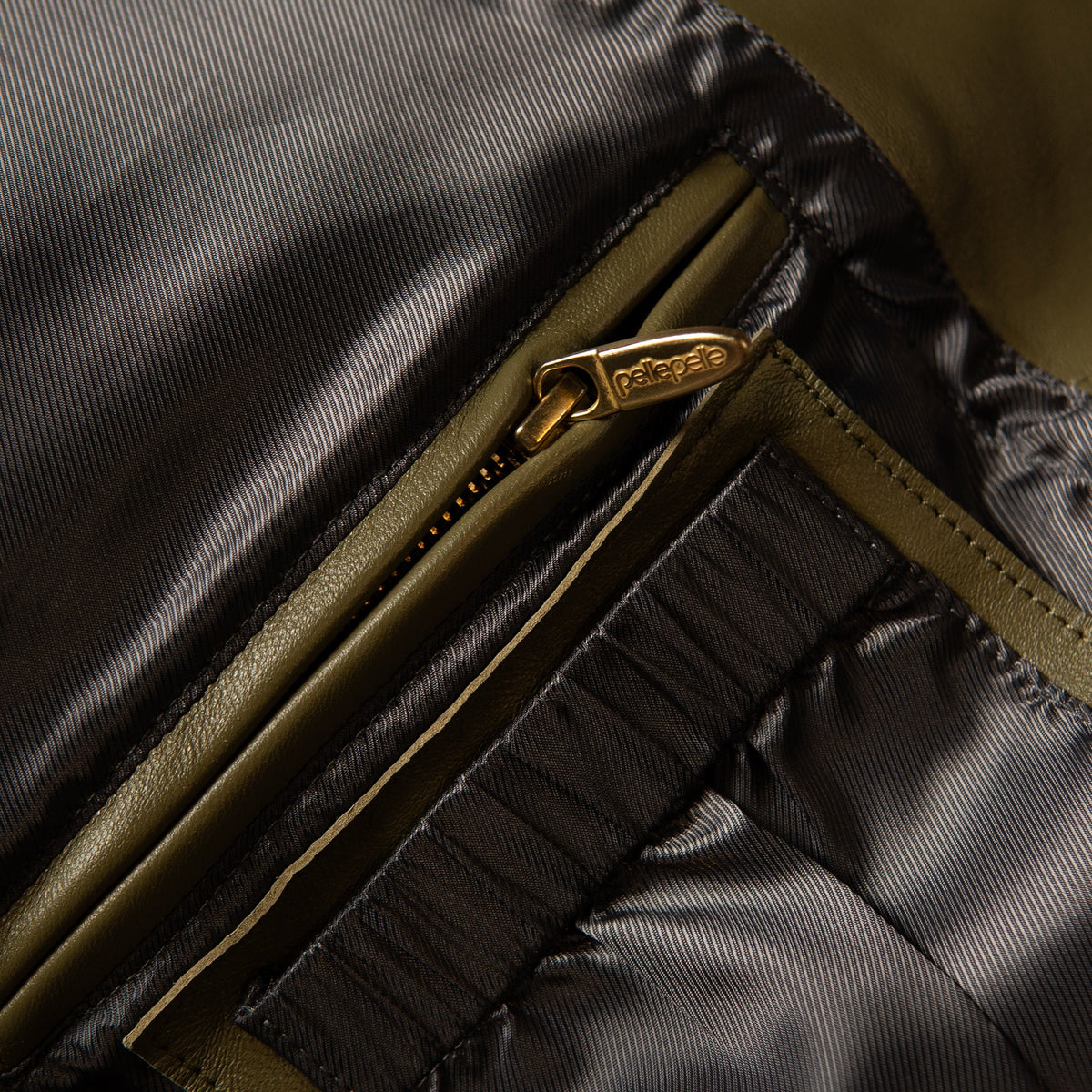Close-up detail of a puffer jacket showing a gold-tone zipper with a 'pelle' pull tab, olive green trim, and dark gray quilted fabric.