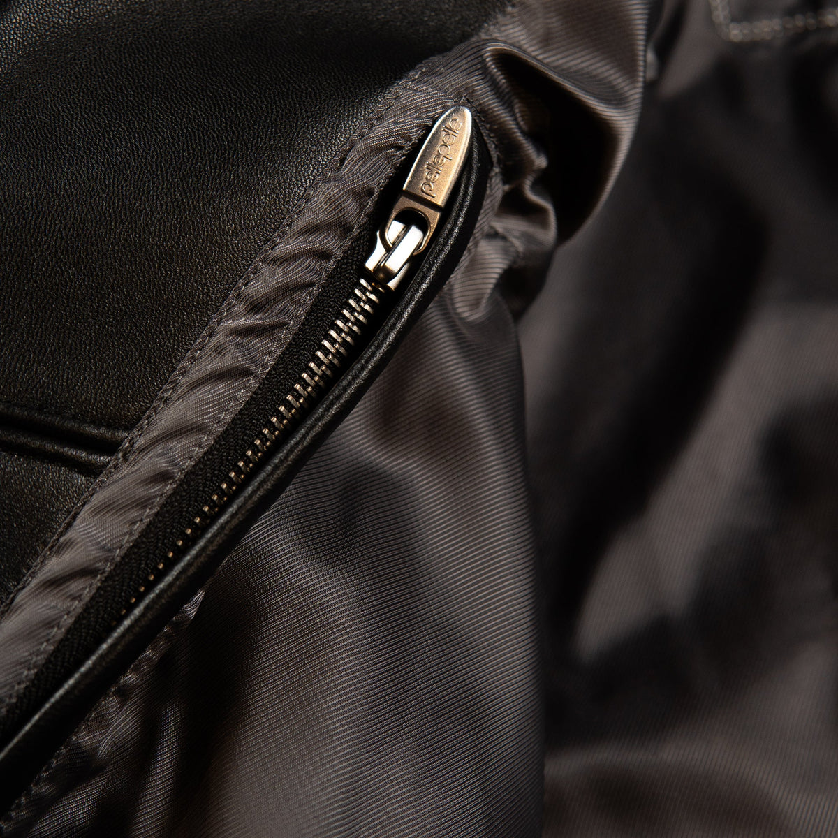 Close-up of a black nylon garment with a silver metal zipper, showing the zipper pull engraved with 'PELLE'.
