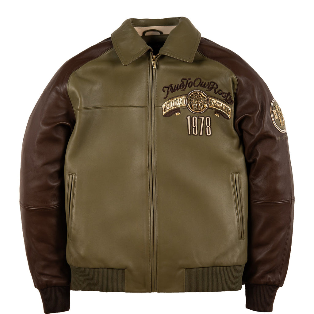 An olive green and dark brown leather jacket with a front zipper, ribbed collar, cuffs, and hem. The left chest features a circular patch with the text 'True To Our Roots' and '1978', and there is a circular patch on the right sleeve.