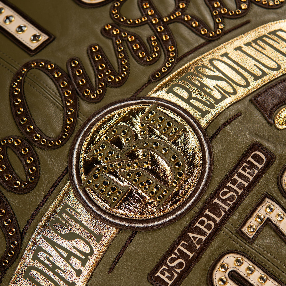 Close-up of an olive green leather jacket featuring metallic gold and bronze embroidered patches with text such as 'RESOLUTION', 'FAST', 'ESTABLISHED', and a central circular patch with a horse and rider motif.
