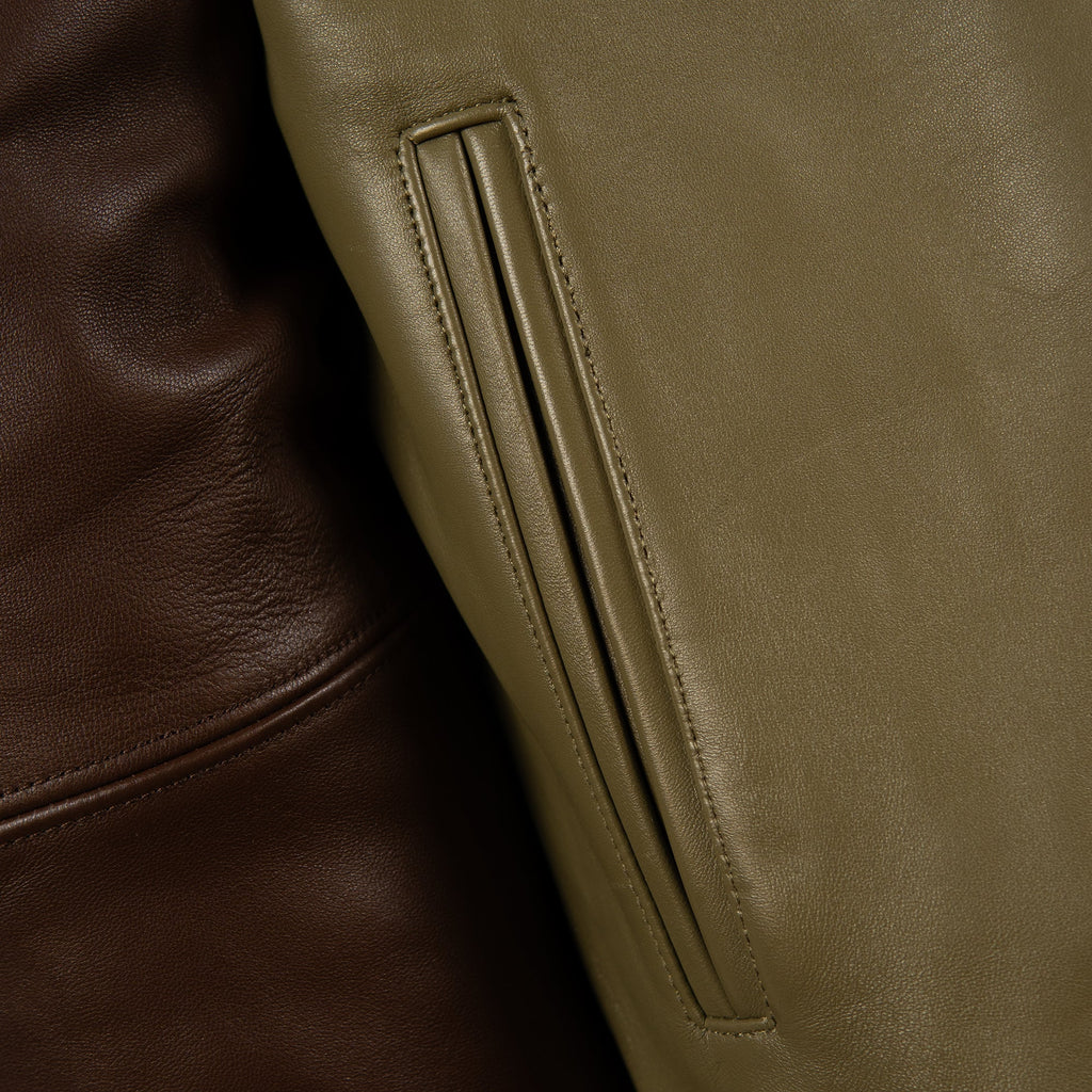 Close-up detail of a leather jacket showing a vertical welt pocket in olive green alongside a darker brown leather section.
