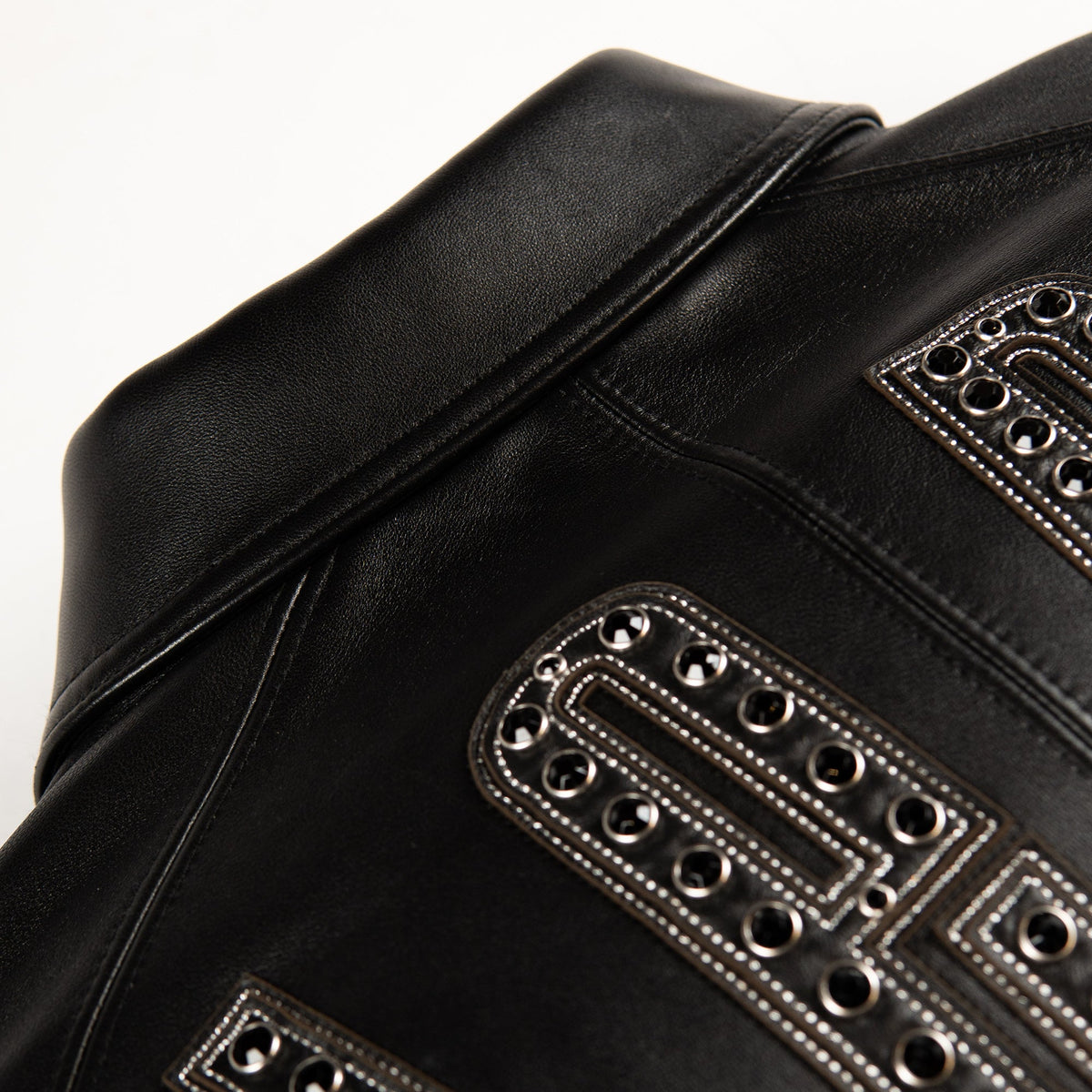 Close-up of a black leather jacket featuring geometric silver-tone metal studs and contrast stitching on a white background.