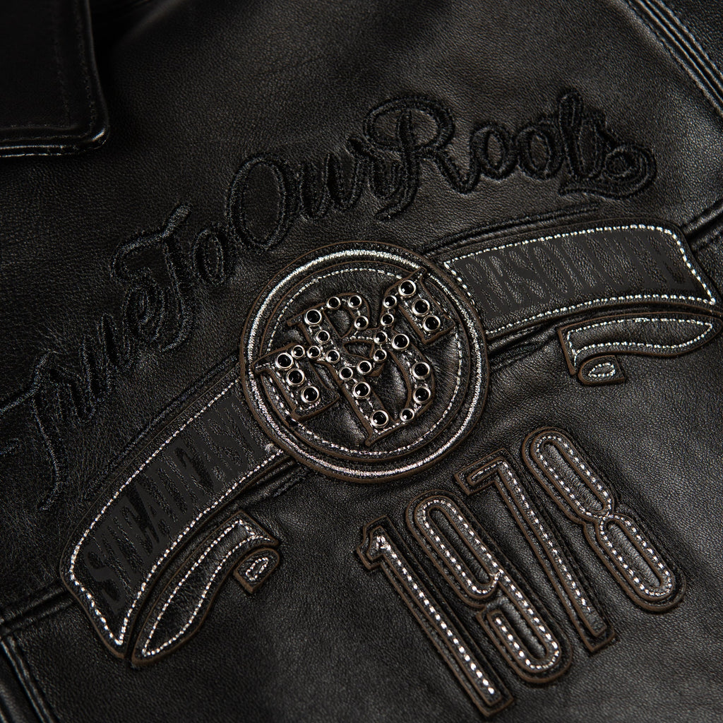 Close-up of a black leather jacket featuring an embroidered circular crest with the letters 'R&R' and the script text 'True To Our Roots' above the number '1978'.