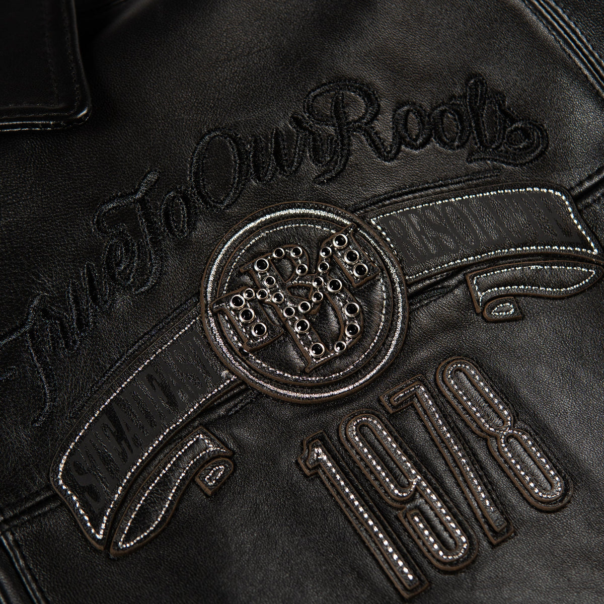 Close-up of a black leather jacket featuring an embroidered circular crest with the letters 'R&R' and the script text 'True To Our Roots' above the number '1978'.