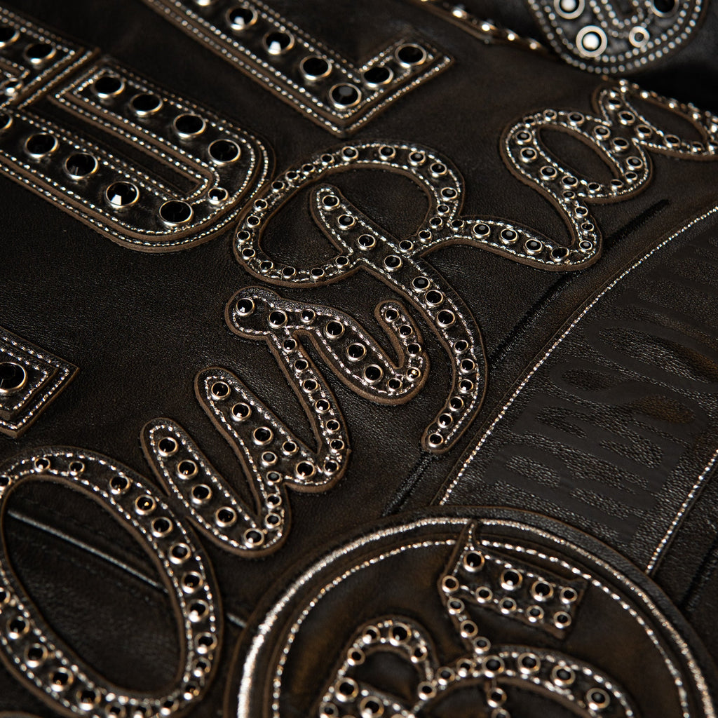 Close-up of a black leather garment featuring large, ornate silver-tone embroidered letters and mirrored rhinestone embellishments.