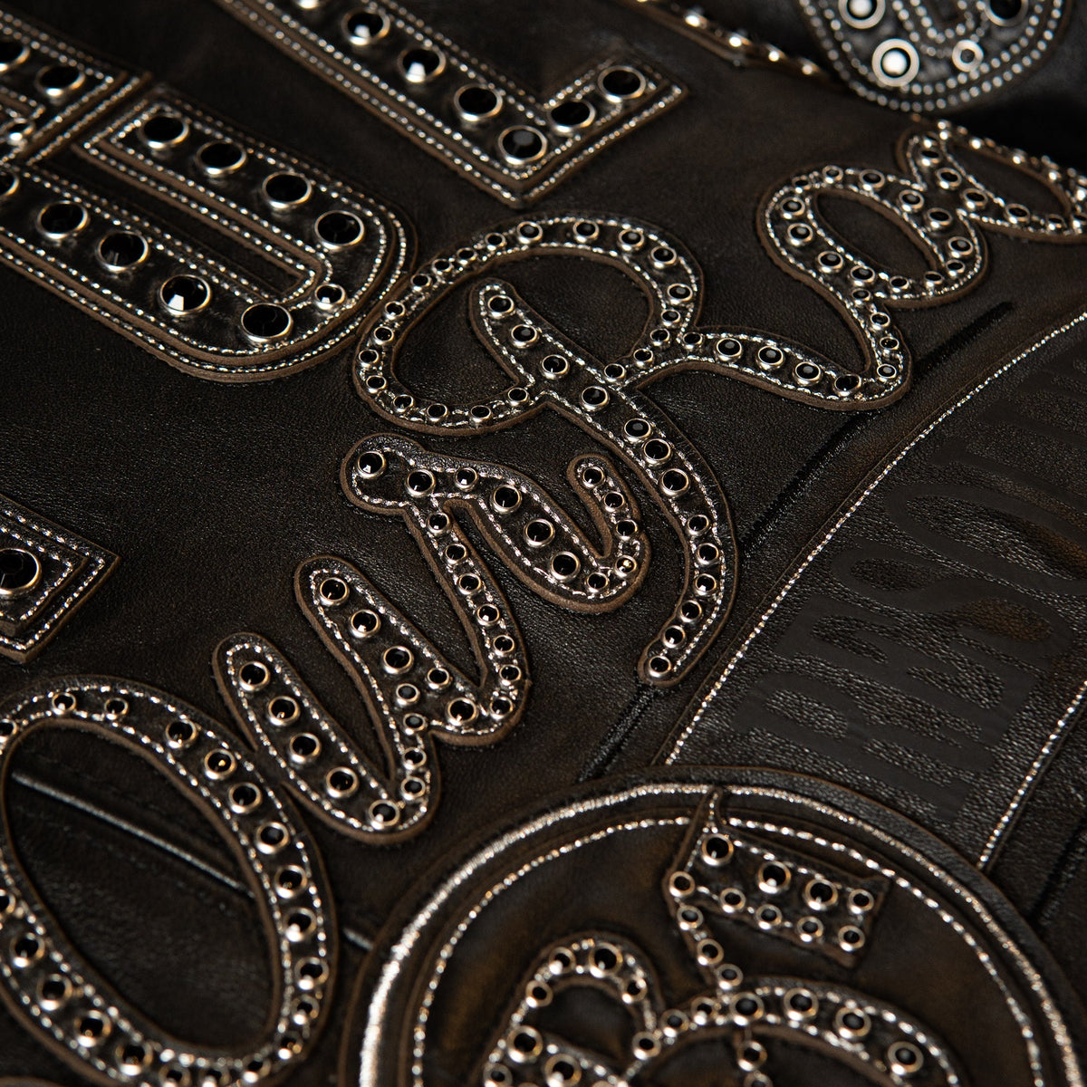 Close-up of a black leather garment featuring large, ornate silver-tone embroidered letters and mirrored rhinestone embellishments.