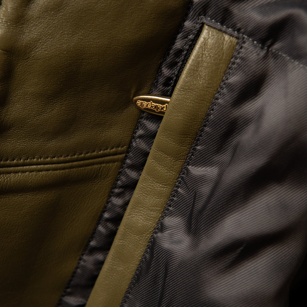 Close-up detail of a jacket showing olive green leather trim, a black nylon interior, and a small gold-tone metal logo badge.